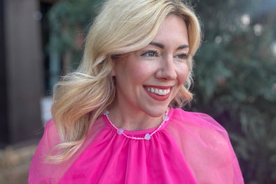 Woman wearing a pink dress with a sheer overlay and pearl accents, smiling outdoors.