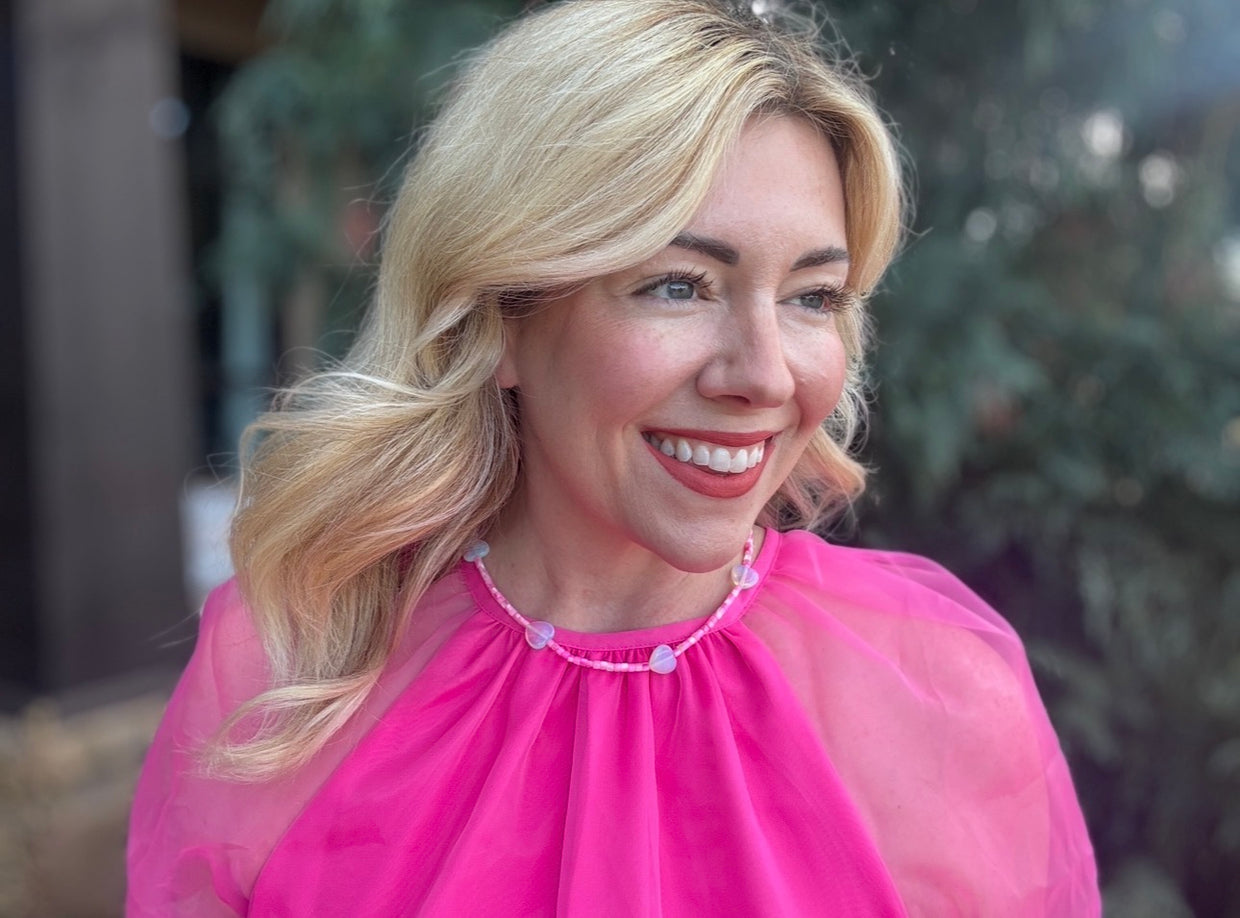 Woman wearing a pink dress with a sheer overlay and pearl accents, smiling outdoors.